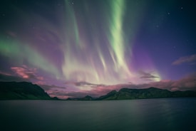 Vivid aurora borealis in the night sky, with green and purple lights dancing above a serene lake. The lights illuminate the surrounding rugged mountains and create reflections on the water below.