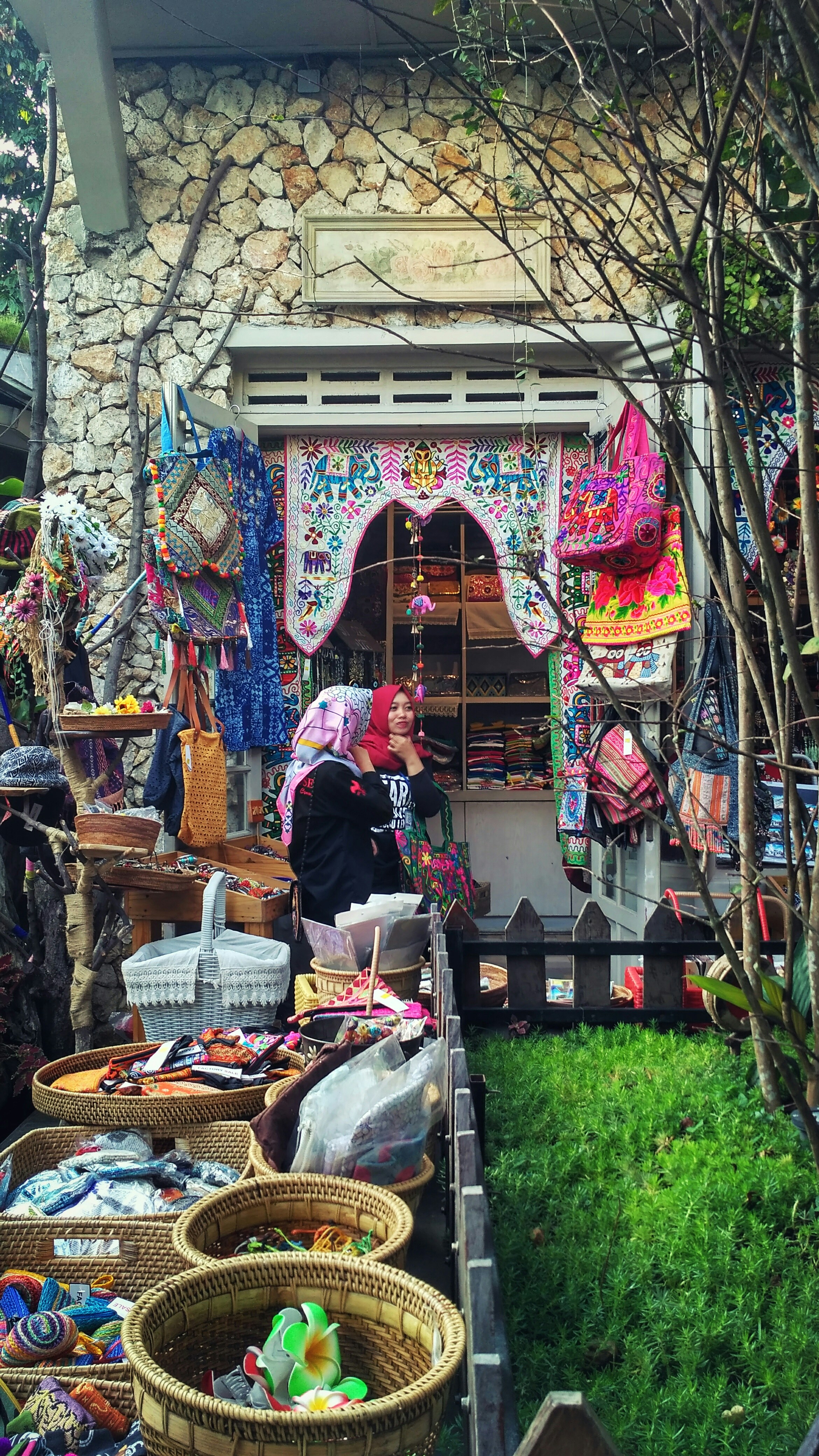 A quaint market or shop display featuring vibrant, colorful textiles and handmade crafts. Baskets are filled with various trinkets, while the surrounding area includes lush greenery, stone walls, and intricate decorations. Two people are visible, seemingly admiring or organizing the goods.