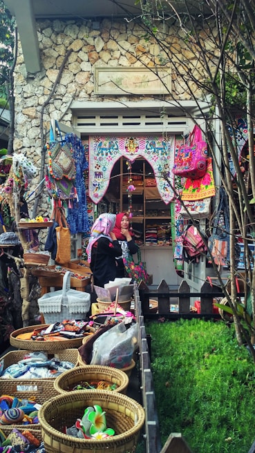 A quaint market or shop display featuring vibrant, colorful textiles and handmade crafts. Baskets are filled with various trinkets, while the surrounding area includes lush greenery, stone walls, and intricate decorations. Two people are visible, seemingly admiring or organizing the goods.