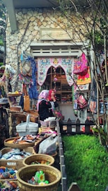 A quaint market or shop display featuring vibrant, colorful textiles and handmade crafts. Baskets are filled with various trinkets, while the surrounding area includes lush greenery, stone walls, and intricate decorations. Two people are visible, seemingly admiring or organizing the goods.