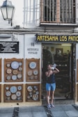 A person stands outside a shop named Artesania Los Patios, capturing photographs with a camera. The shop's exterior features decorative ceramic plates displayed on the walls, with intricate designs. The building has a traditional appearance with a white facade and black iron grilles on the windows. A lamp is mounted on the wall beside the shop entrance.