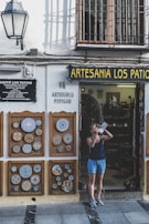 A person stands outside a shop named Artesania Los Patios, capturing photographs with a camera. The shop's exterior features decorative ceramic plates displayed on the walls, with intricate designs. The building has a traditional appearance with a white facade and black iron grilles on the windows. A lamp is mounted on the wall beside the shop entrance.