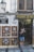 A person stands outside a shop named Artesania Los Patios, capturing photographs with a camera. The shop's exterior features decorative ceramic plates displayed on the walls, with intricate designs. The building has a traditional appearance with a white facade and black iron grilles on the windows. A lamp is mounted on the wall beside the shop entrance.