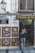 A person stands outside a shop named Artesania Los Patios, capturing photographs with a camera. The shop's exterior features decorative ceramic plates displayed on the walls, with intricate designs. The building has a traditional appearance with a white facade and black iron grilles on the windows. A lamp is mounted on the wall beside the shop entrance.