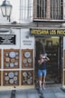 A person stands outside a shop named Artesania Los Patios, capturing photographs with a camera. The shop's exterior features decorative ceramic plates displayed on the walls, with intricate designs. The building has a traditional appearance with a white facade and black iron grilles on the windows. A lamp is mounted on the wall beside the shop entrance.