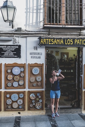 A person stands outside a shop named Artesania Los Patios, capturing photographs with a camera. The shop's exterior features decorative ceramic plates displayed on the walls, with intricate designs. The building has a traditional appearance with a white facade and black iron grilles on the windows. A lamp is mounted on the wall beside the shop entrance.