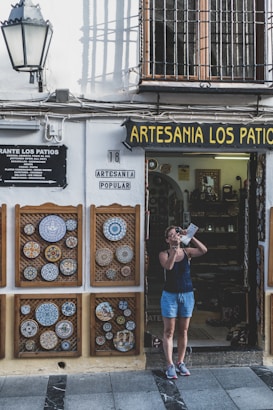 A person stands outside a shop named Artesania Los Patios, capturing photographs with a camera. The shop's exterior features decorative ceramic plates displayed on the walls, with intricate designs. The building has a traditional appearance with a white facade and black iron grilles on the windows. A lamp is mounted on the wall beside the shop entrance.