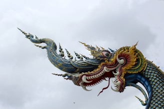 An intricately designed dragon sculpture with elaborate detailing and vibrant colors. Its face is adorned with curling tendrils and sharp teeth, while its body scales are artistically rendered. The dragon's head is positioned against a cloudy sky, showcasing its mythological features.