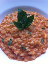 A bowl of creamy risotto with a rich orange color, topped with fresh basil leaves. The grains are well-cooked and evenly coated with sauce, with flecks of green herbs adding contrast.