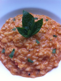 A bowl of creamy risotto with a rich orange color, topped with fresh basil leaves. The grains are well-cooked and evenly coated with sauce, with flecks of green herbs adding contrast.