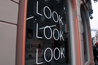 A storefront window with a neon sign displaying the word 'LOOK' repeated three times in vertical alignment. The interior of the store is visible through the glass, showing shelves with products and wall decor. The exterior features a pinkish frame and partial view of the street outside.