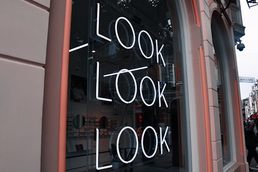 A storefront window with a neon sign displaying the word 'LOOK' repeated three times in vertical alignment. The interior of the store is visible through the glass, showing shelves with products and wall decor. The exterior features a pinkish frame and partial view of the street outside.