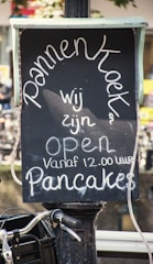 A chalkboard sign displays the word 'Pannenkoeken' prominently. Beneath it, the text reads 'wij zijn open vanaf 12.00 uur Pancakes', indicating an establishment selling pancakes and their opening hours. The sign is set against an outdoor backdrop, suggesting a street or market setting.