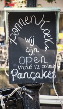 A chalkboard sign displays the word 'Pannenkoeken' prominently. Beneath it, the text reads 'wij zijn open vanaf 12.00 uur Pancakes', indicating an establishment selling pancakes and their opening hours. The sign is set against an outdoor backdrop, suggesting a street or market setting.