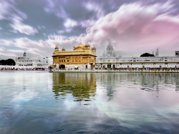 A stunning view of a golden building, likely the Golden Temple, standing majestically by a calm water body. The structure is surrounded by white architecture, and the sky is filled with dramatic pink and purple clouds. There is a reflection of the buildings and sky in the water, creating a serene and harmonious scene.