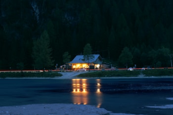 A cozy cabin with warm lights emanates a welcoming glow amidst a dark and dense forest. Reflections of the cabin lights shimmer on the calm waters of a nearby lake. A few cars are parked nearby, visible due to the trail of their headlights lining the road. Tall trees surround the area, silhouetted against the night sky.