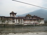 An impressive historic structure with traditional architecture, featuring a large building with multiple tiers and ornate wooden facades. The structure is set against a backdrop of misty mountains and bordered by a river with visible stones and banks.