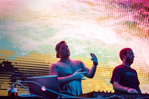 Two individuals are performing at what appears to be a DJ set. The person on the left is engaged with the music equipment, wearing a backward cap and gesturing with their hands. The other individual on the right is focused on the equipment, wearing a t-shirt. A colorful, abstract LED screen is in the background, adding a dynamic and lively atmosphere.