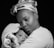 A woman holds a baby closely, wrapped in a soft, textured blanket. The woman has a gentle expression and wears a patterned head wrap. The image is black and white, emphasizing the emotional connection between the subjects against a dark background.