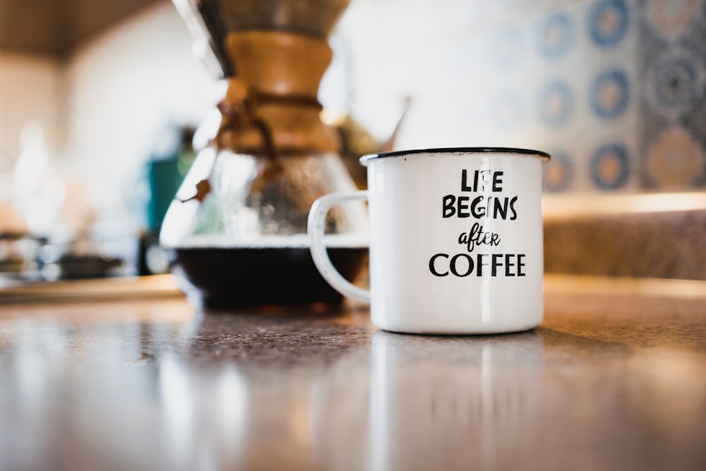 white and black life begins after coffee printed enamel cup beside 1/4 black liquid-filled glass flask on brown wooden panel