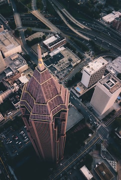 An aerial view of a cityscape showcasing a tall, uniquely designed building with angular structures and a pointed roof. Surrounding the skyscraper are several other buildings, roads, and a network of highway overpasses. The urban setting is densely packed with a mix of commercial and office buildings.