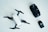A small white drone with four propellers is placed on a light surface. Nearby, there is a black remote control with various buttons and joysticks. To the right of the remote, there is a battery pack with a grid symbol on top.