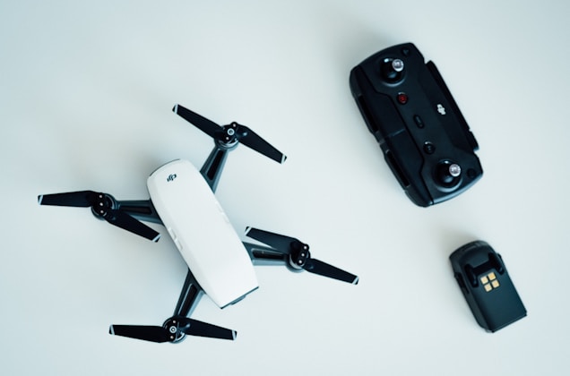 A small white drone with four propellers is placed on a light surface. Nearby, there is a black remote control with various buttons and joysticks. To the right of the remote, there is a battery pack with a grid symbol on top.