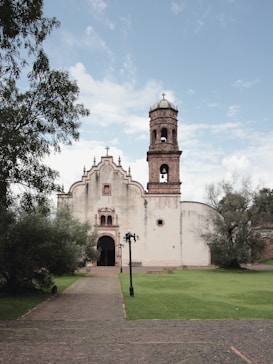 A historic stone church with a baroque architectural style and a prominent bell tower stands surrounded by lush greenery. The path leading to the entrance is paved with bricks and flanked by well-maintained grass and mature trees. The sky is mostly clear with a few clouds.