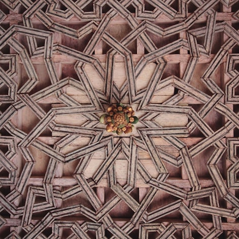 Intricate geometric wooden pattern featuring interlocking star and polygon shapes with a detailed floral motif at the center. The design showcases a layered, three-dimensional effect created by the overlapping wood pieces.