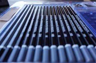 A set of graphite pencils arranged neatly in a case. The pencils are aligned parallel to each other, with their labels visible and the sharp ends pointed towards the right. The case includes a plastic cover and a cardboard flap on the side.