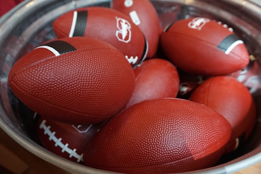 A pile of red American footballs is placed inside a large metallic container. Each ball has a small black stripe near the tips and prominent white stitching. The textured surface of the footballs is visible, indicating they might be new and unused.