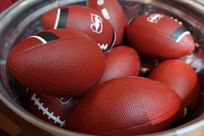 A pile of red American footballs is placed inside a large metallic container. Each ball has a small black stripe near the tips and prominent white stitching. The textured surface of the footballs is visible, indicating they might be new and unused.