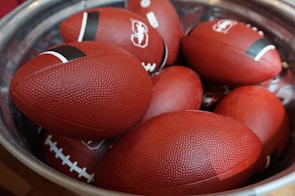 A pile of red American footballs is placed inside a large metallic container. Each ball has a small black stripe near the tips and prominent white stitching. The textured surface of the footballs is visible, indicating they might be new and unused.