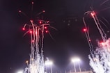 Bright fireworks with red lights and trailing smoke explode against a dark night sky above stadium lights.