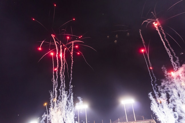 Bright fireworks with red lights and trailing smoke explode against a dark night sky above stadium lights.