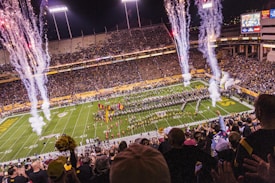 A large stadium filled with spectators watching a marching band and cheerleaders performing on the football field. Fireworks are being launched in the air, creating a festive and vibrant atmosphere. The stands are crowded, indicating a major sporting event or celebration, and there is a large scoreboard visible in the background.