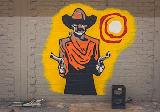 A colorful mural on a brick wall depicts a cowboy wearing a wide-brimmed hat and draped clothing, holding two pistols. The background features a bright yellow halo with a circular design in red and white. Below the mural, there is a black crate and some metal frames on the ground.