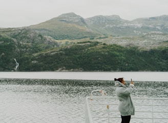 A person wearing a gray coat is taking a photo with a smartphone while standing on a boat. The background features a scenic view of a fjord surrounded by towering, green mountainous landscapes under a cloudy sky.