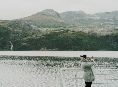 A person wearing a gray coat is taking a photo with a smartphone while standing on a boat. The background features a scenic view of a fjord surrounded by towering, green mountainous landscapes under a cloudy sky.