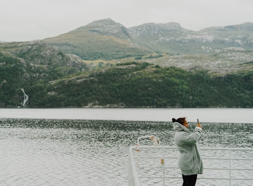 A person wearing a gray coat is taking a photo with a smartphone while standing on a boat. The background features a scenic view of a fjord surrounded by towering, green mountainous landscapes under a cloudy sky.