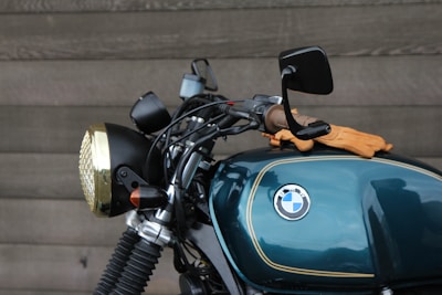 A close-up view of a motorcycle featuring a dark teal fuel tank with a BMW logo, alongside handlebars and a headlight with a brass-colored trim. A pair of brown leather gloves is resting on the tank. The background is a textured wooden wall.