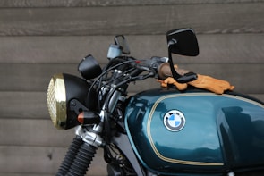 A close-up view of a motorcycle featuring a dark teal fuel tank with a BMW logo, alongside handlebars and a headlight with a brass-colored trim. A pair of brown leather gloves is resting on the tank. The background is a textured wooden wall.