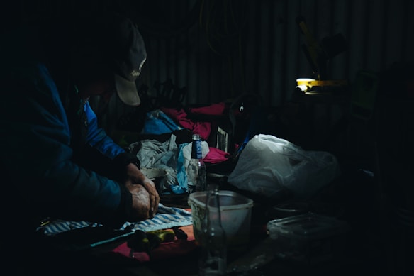 A dimly lit workspace in which a person is working with their hands on a striped cloth. Surrounding the individual are various items including a plastic bottle, a transparent container, and scattered clothes or fabric in different colors, with some illuminated by a focused light source.