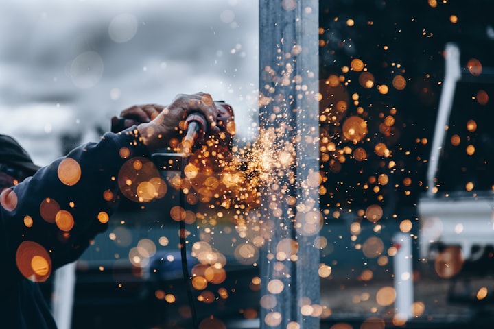 A construction worker using an angle grinder, sending sparks flying as its being used on a girder.