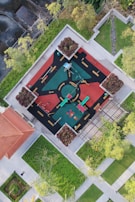 An aerial view of a vibrant playground surrounded by greenery. The playground features colorful equipment such as slides and swings, set on a patterned surface with road-like markings. Trees and landscaping surround the playground, providing a natural border.