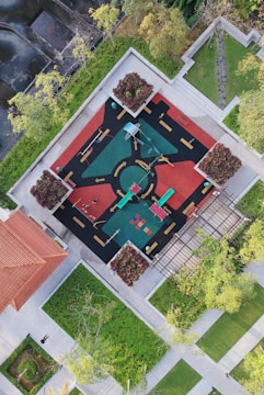An aerial view of a vibrant playground surrounded by greenery. The playground features colorful equipment such as slides and swings, set on a patterned surface with road-like markings. Trees and landscaping surround the playground, providing a natural border.