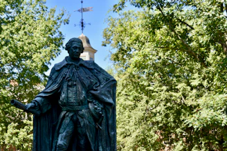 A bronze statue of a historical figure stands amidst lush green trees. The figure is dressed in 18th-century attire, holding a scroll in one hand and an hourglass in the other, with an authoritative pose. In the background, a chapel or similar structure with a weather vane on the roof is visible against a clear blue sky.