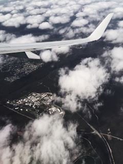 An aerial view featuring a section of a plane wing slicing through the upper part of the frame, looking down on partially cloudy skies. Below, a patchwork of industrial buildings and lush green areas is visible, interspersed with roads and surrounded by expanses of forest.