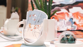 A brightly lit table setting features a white mug filled with colorful business cards, alongside a small white teapot. In the background, there is a piece of abstract art with bold strokes of orange, blue, and white. A succulent plant in a white pot and a decorative glass orb are also present on the table.