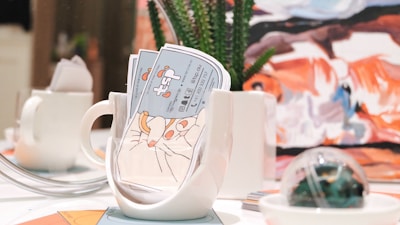 A brightly lit table setting features a white mug filled with colorful business cards, alongside a small white teapot. In the background, there is a piece of abstract art with bold strokes of orange, blue, and white. A succulent plant in a white pot and a decorative glass orb are also present on the table.